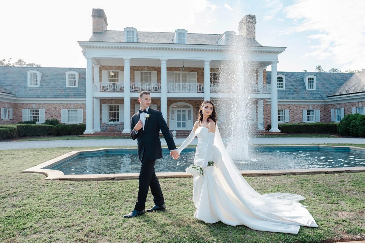 Bride and groom walking past a fountain in front of a white columned estate