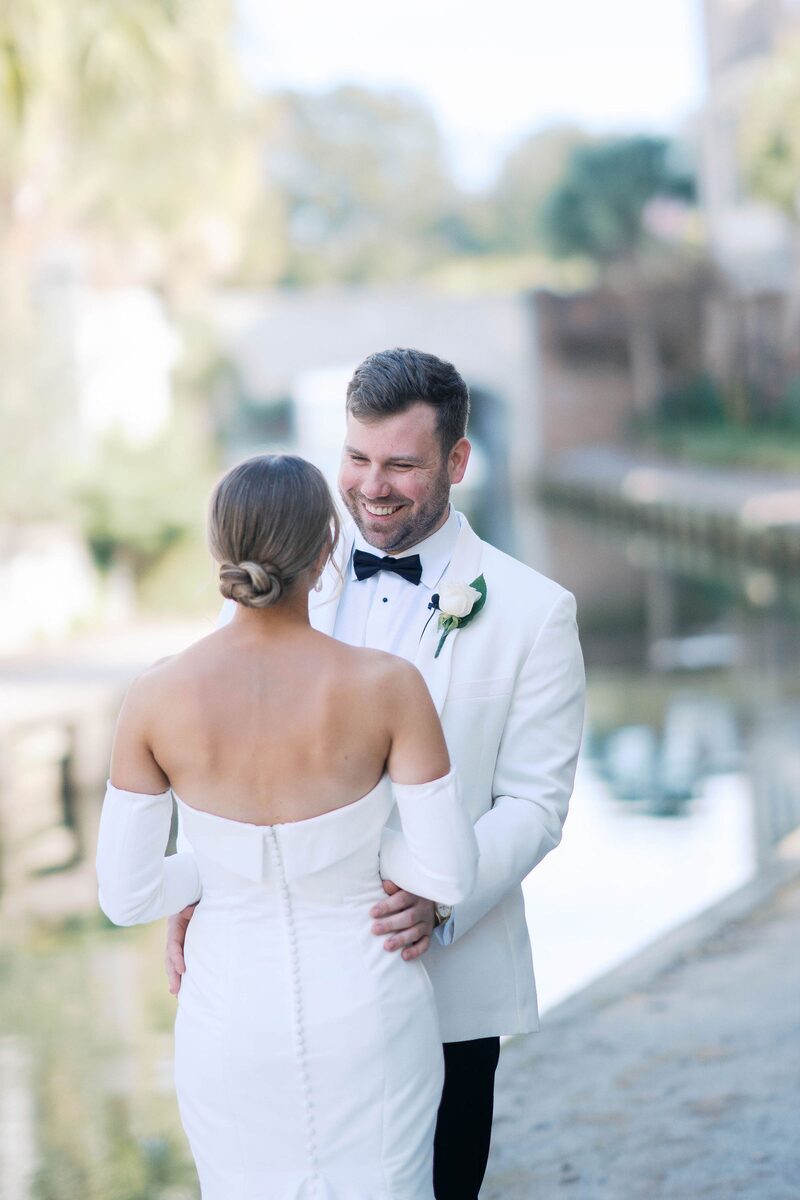 The groom in a white tuxedo with his bride by the water