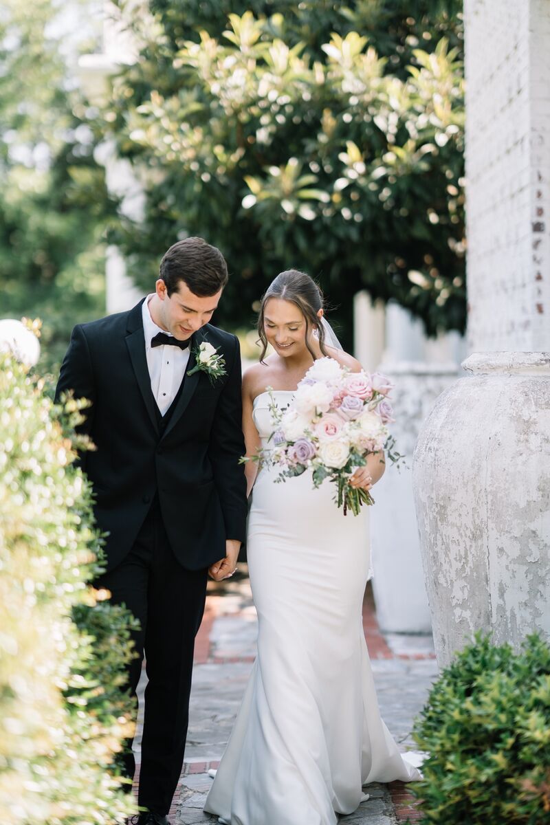 Bride and groom walking along a garden path, her bouquet of pink and white flowers