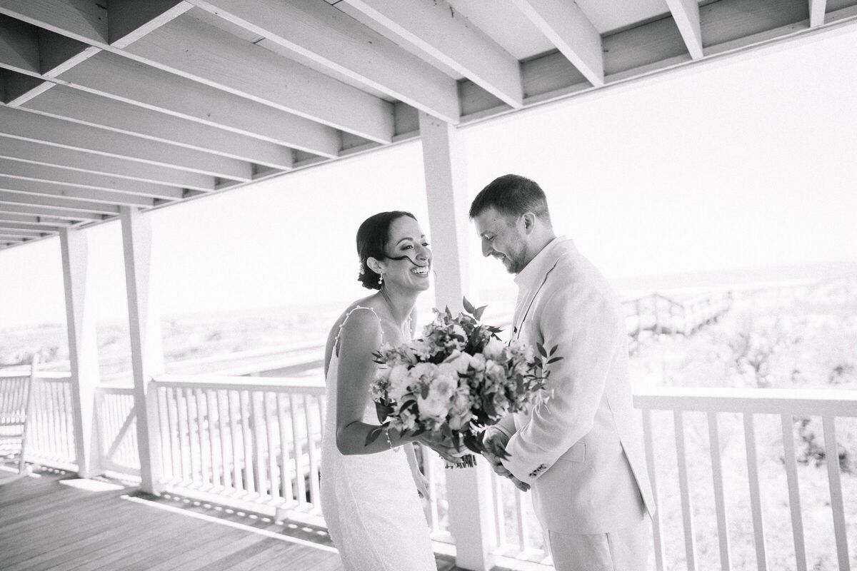 Bride and groom laughing on a white wraparound porch, her bouquet in hand