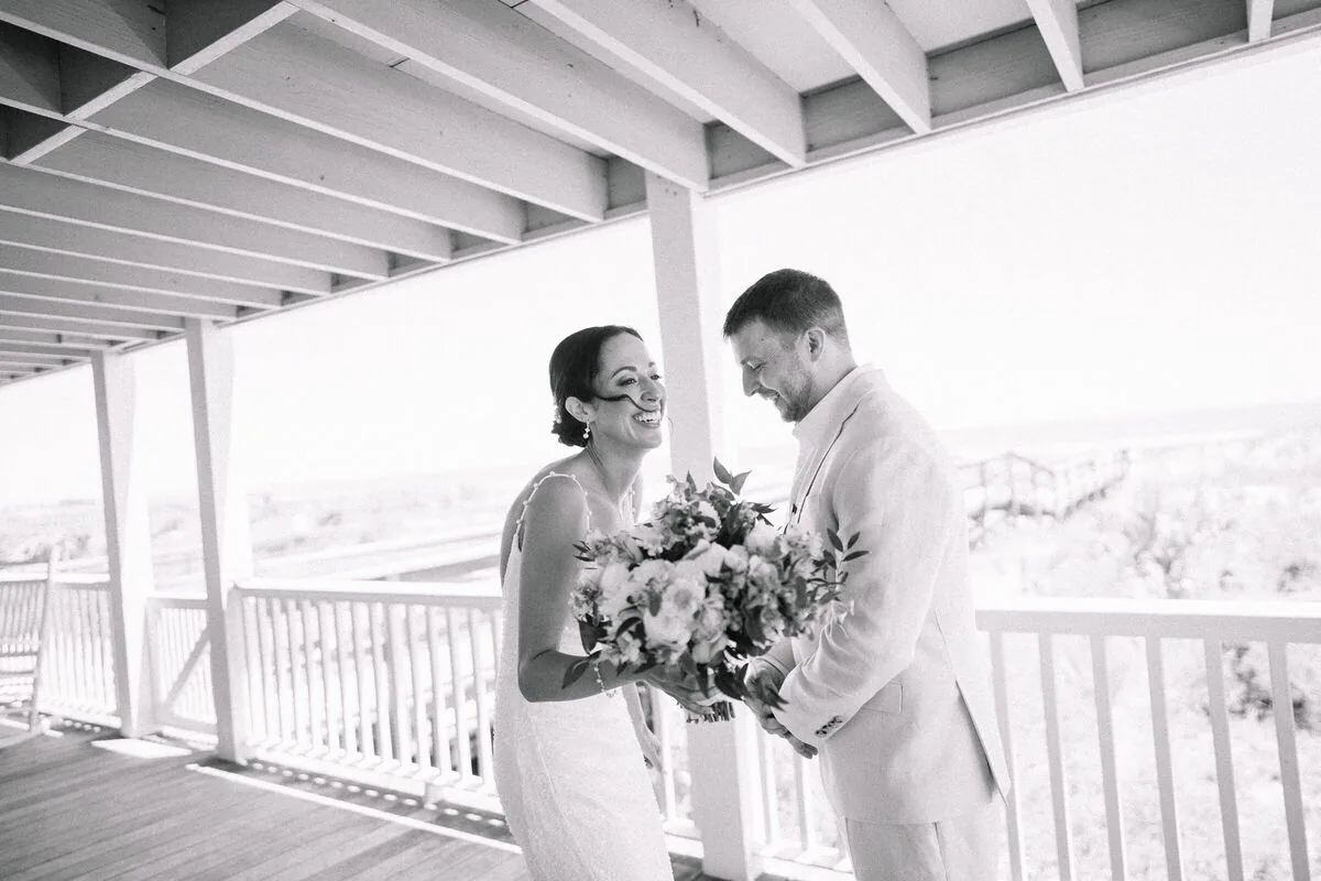 Bride and groom laughing on a white wraparound porch, her bouquet in hand