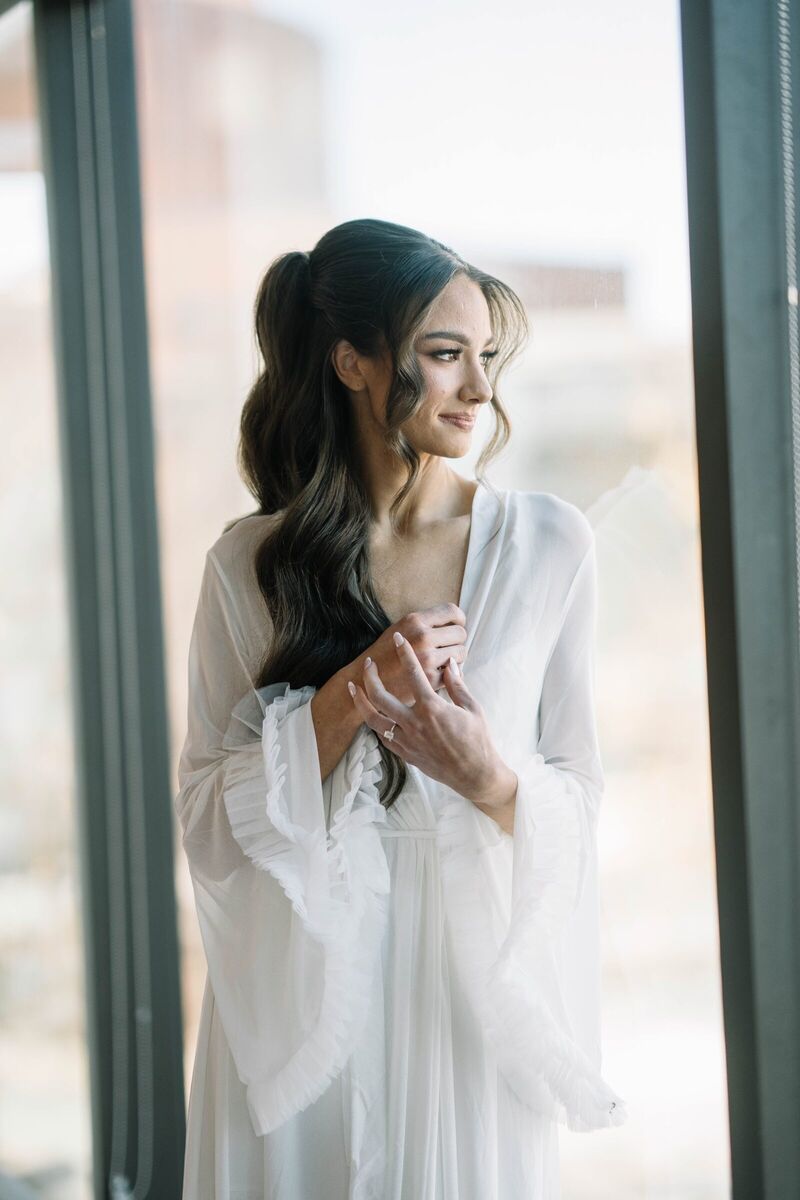 Bride in a white robe by a sunlit window on her wedding morning