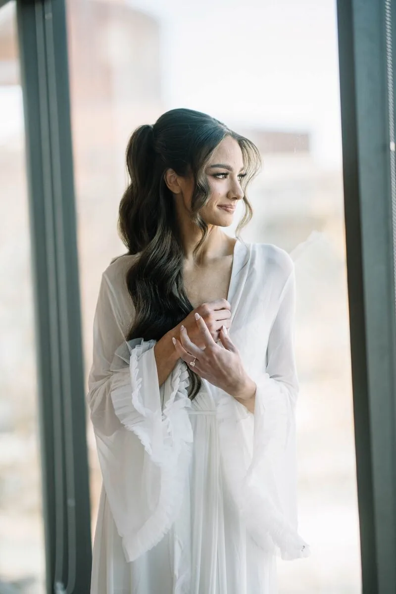 Bride in a white robe by a sunlit window on her wedding morning
