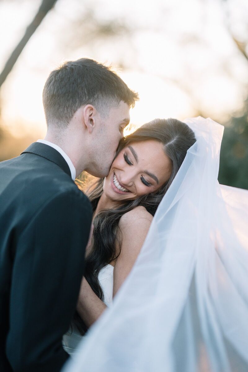 Groom kissing his smiling bride on the cheek at golden hour, her veil catching the light