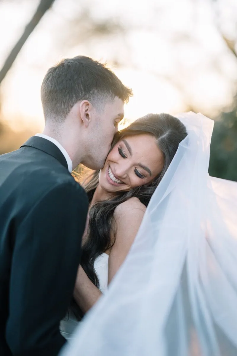 Groom kissing his smiling bride on the cheek at golden hour, her veil catching the light